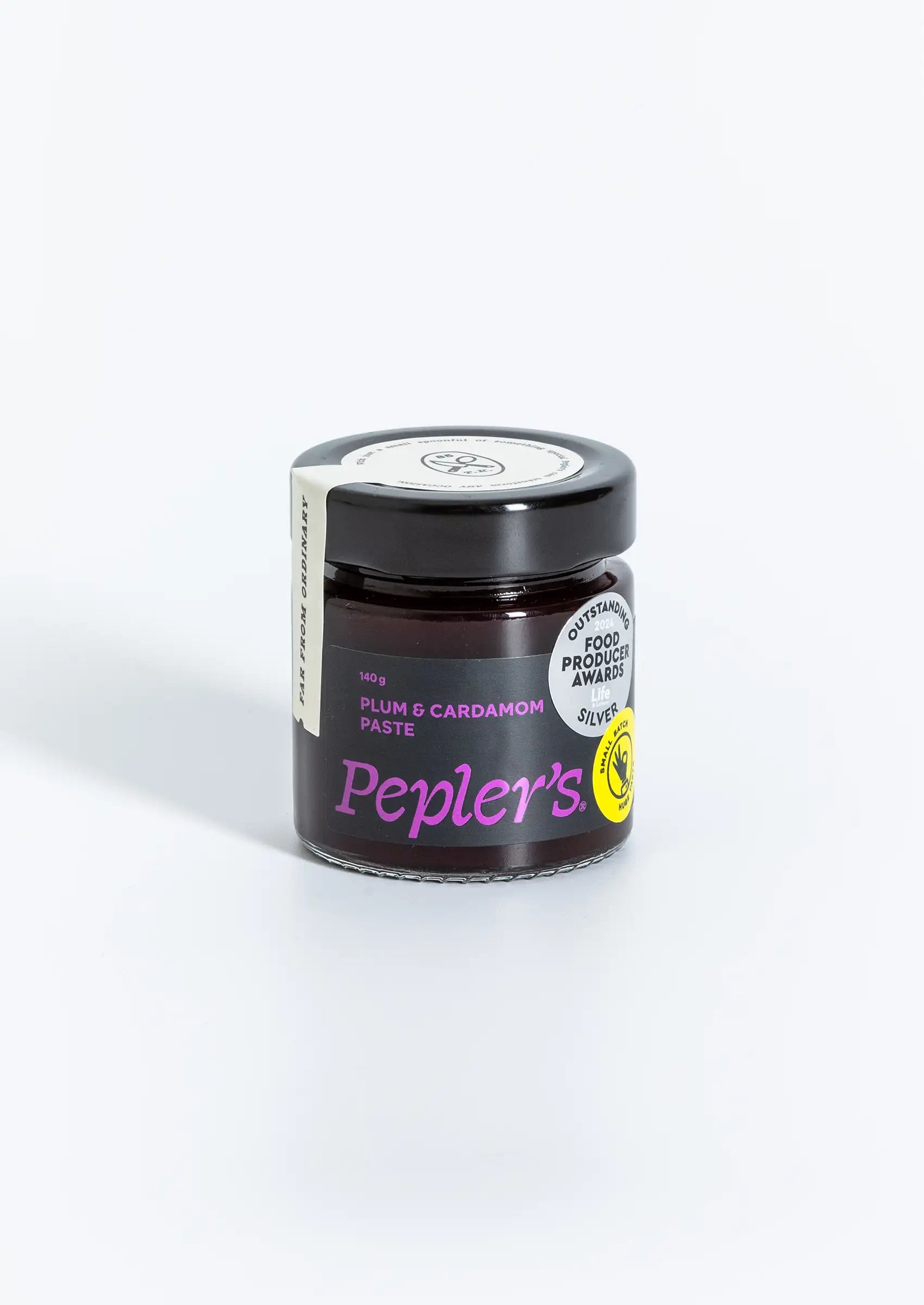 A clear glass jar containing plum and cardamom paste, featuring a black lid. The label is predominantly black with bright purple text stating "PLUM & CARDAMOM PASTE" and includes the brand name "Pepler's." The jar size is indicated as 140 grams, and a small circular logo in yellow suggests a focus on quality or natural ingredients. The background is plain and light-coloured, highlighting the jar.