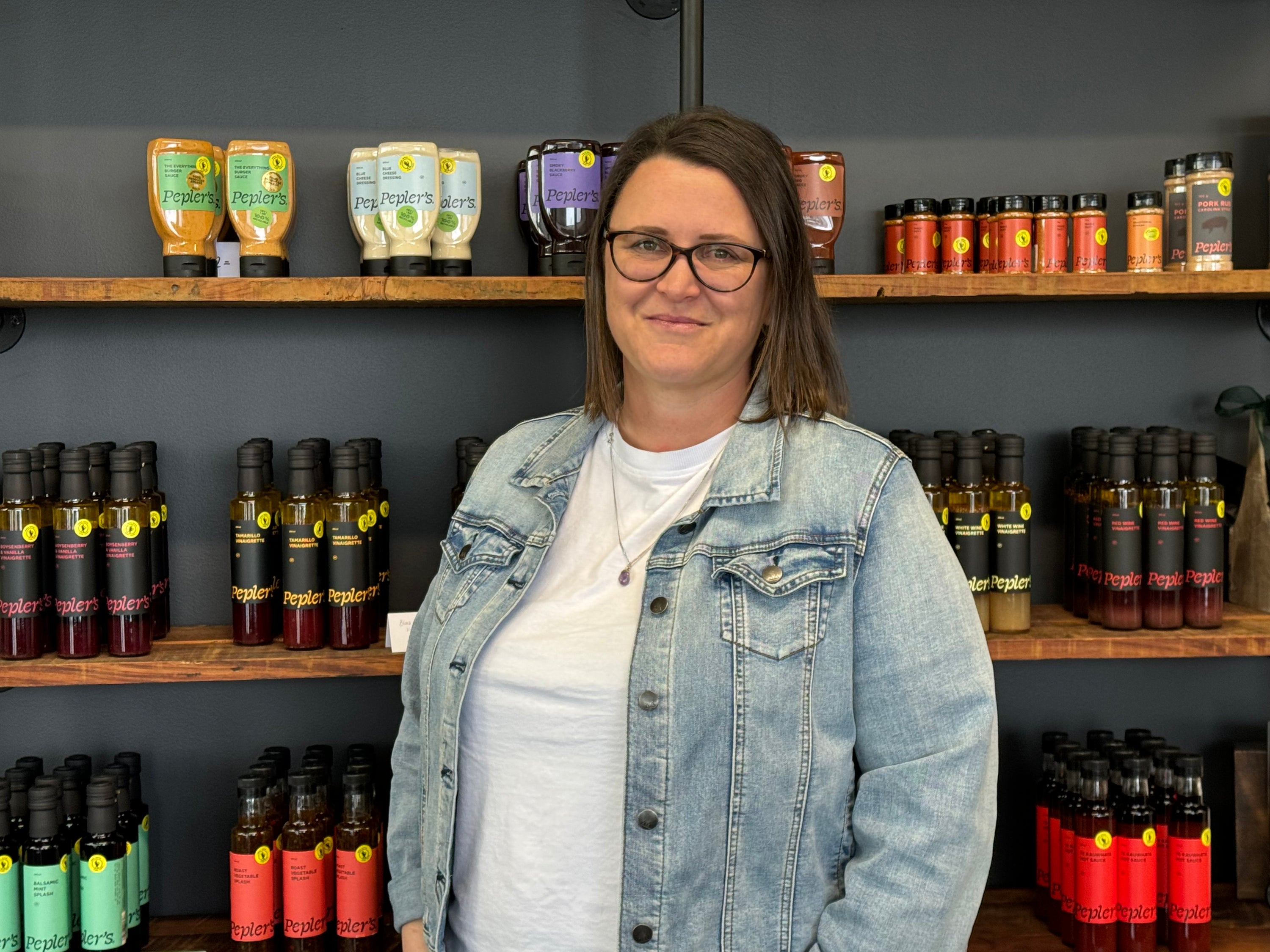 A woman, Anna, with shoulder-length brown hair and glasses stands in front of a shelving unit filled with various bottles and jars of Pepler's products. The shelves display a range of sauces, marinades, and spices, featuring colourful labels in shades of green, yellow, red, and black, all arranged neatly against a dark wall. The woman is wearing a light-coloured t-shirt and a denim jacket, smiling at the camera.