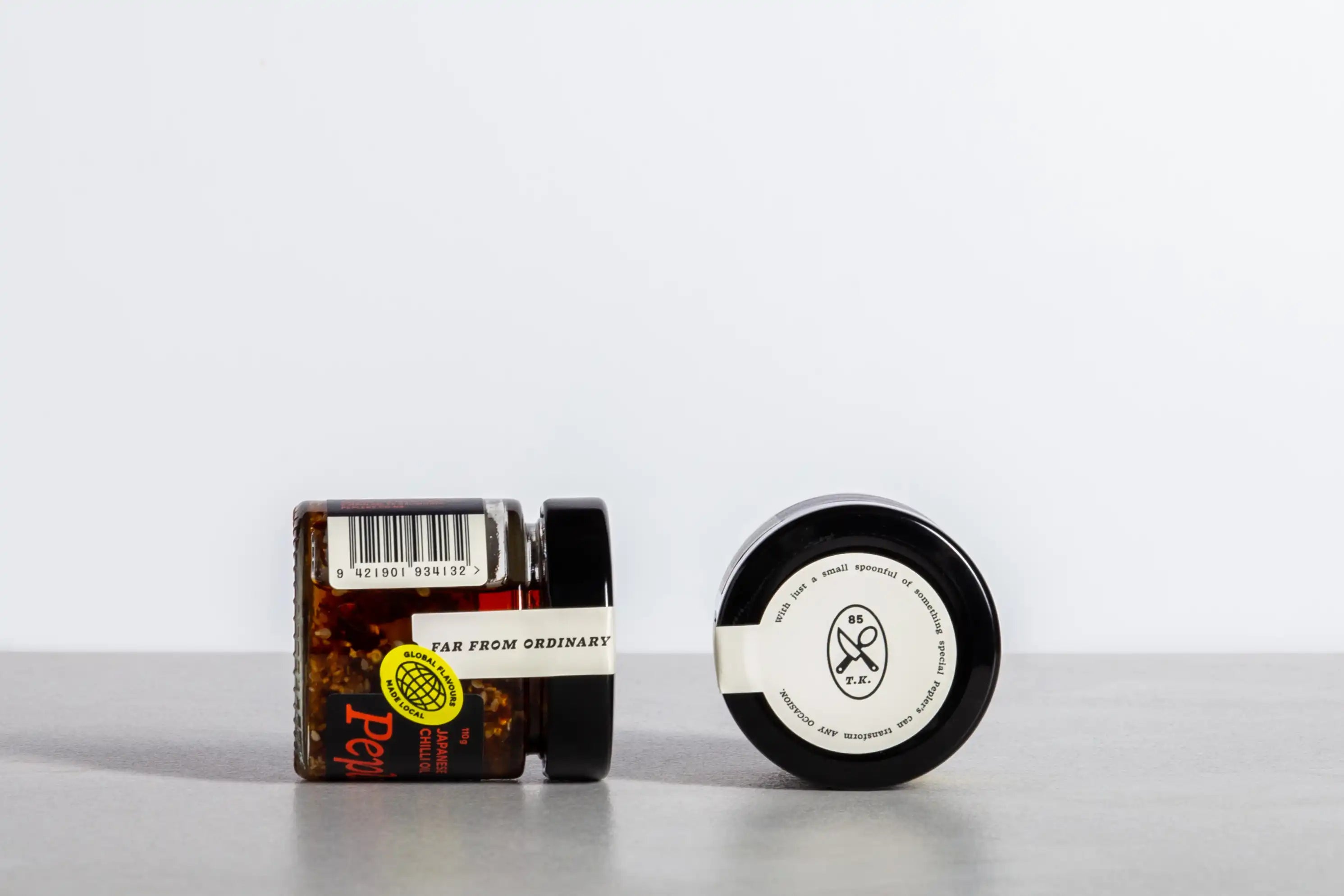 Two jars of Pepler's Chilli Oil are displayed on a gray countertop, with one jar slightly tilted. The first jar features a vibrant label with the words "Far From Ordinary" and has a barcode visible. The second jar, which is black and round, displays an emblem and text on its lid, suggesting it contains a gourmet seasoning or condiment. The background is minimalistic, emphasising the jars.