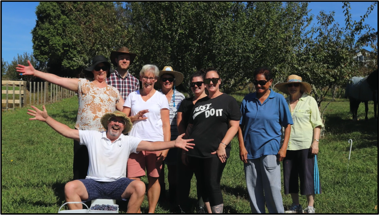 A group of nine people is gathered outdoors on a sunny day, smiling and posing for the camera. They are surrounded by lush greenery and trees, suggesting a rural or agricultural setting. The individuals are wearing summer clothing and hats for sun protection. One man in shorts is sitting on a bucket filled with blueberries, while others stand around him, some with their hands raised in a welcoming gesture.