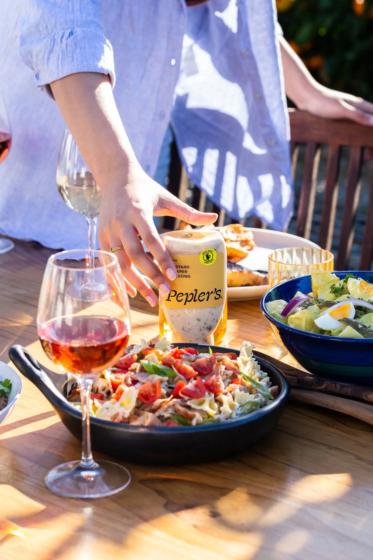 A table set for a meal featuring a hand reaching for a bottle of Pepler's mustard dressing. In the foreground, there is a black dish filled with pasta, cherry tomatoes, and green onions. Nearby, two glasses of rosé wine are visible, along with a colourful bowl of salad topped with hard-boiled eggs. The setting is bright and sunny, with a wooden table and background foliage.