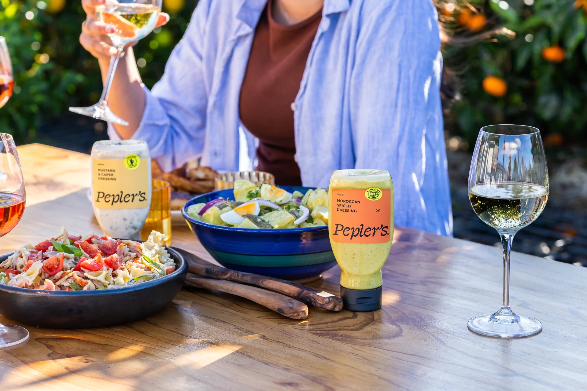 A beautifully arranged outdoor dining table featuring a variety of dishes and drinks. In the foreground, a bowl of pasta salad topped with tomatoes and greens, alongside a colourful salad in a blue bowl. Two bottles of Pepler's dressings, one labeled "Moroccan Spiced Dressing" and the other "Mustard & Capers Dressing," are prominently displayed. Glasses of white wine and rosé are on the table, with a hand holding a glass. The background is filled with vibrant greenery and orange trees.