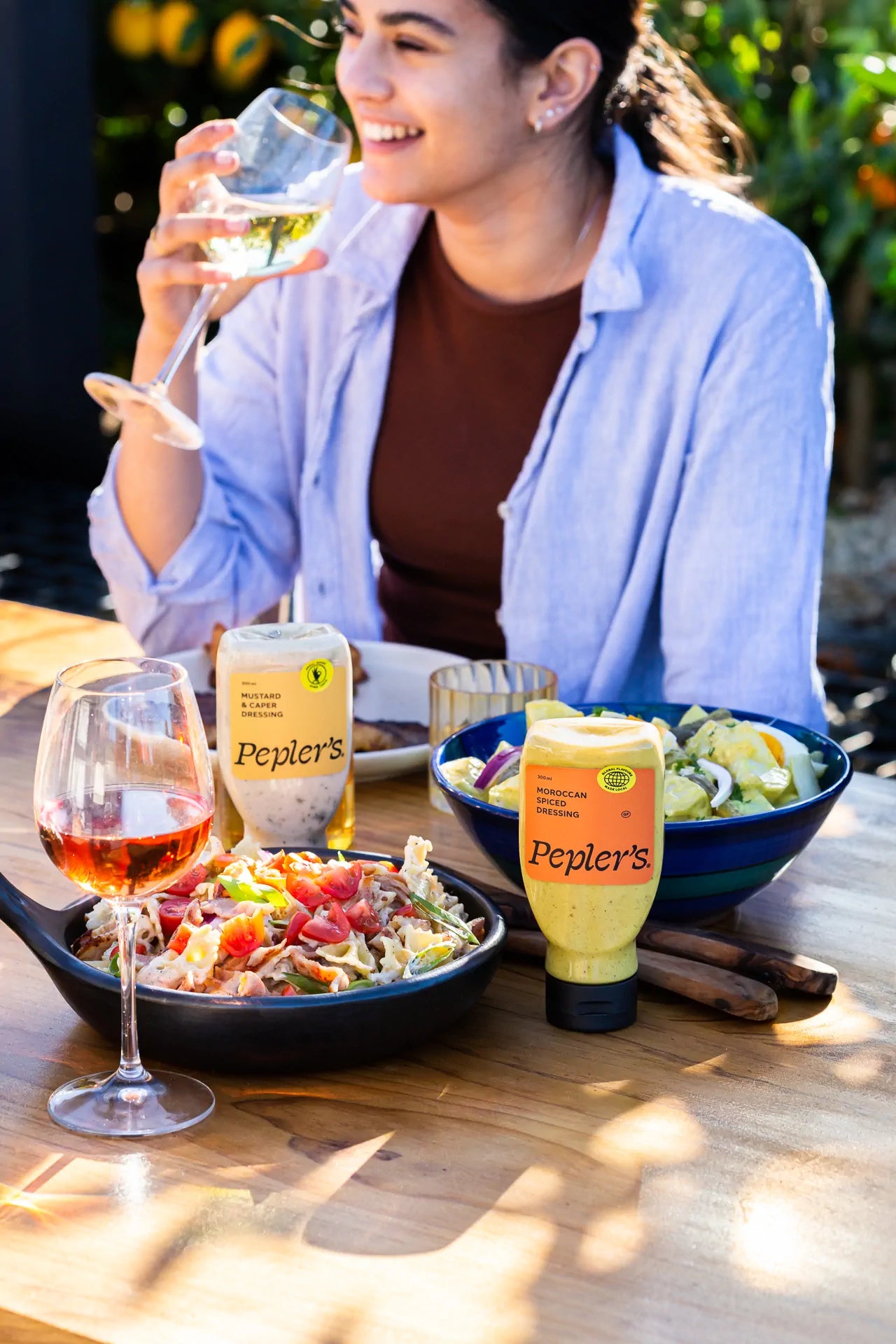 A young woman enjoys a drink while seated at a wooden table, smiling and holding a wine glass filled with a light beverage. In front of her are a variety of dishes, including a black bowl of pasta salad topped with tomatoes, and a blue bowl filled with mixed vegetables. Two bottles of Pepler's salad dressings—one labeled "Mustard & Caper" and the other "Moroccan Spiced"—are prominently placed beside the food. The scene is set outdoors, with green foliage in the background and warm natural lighting.