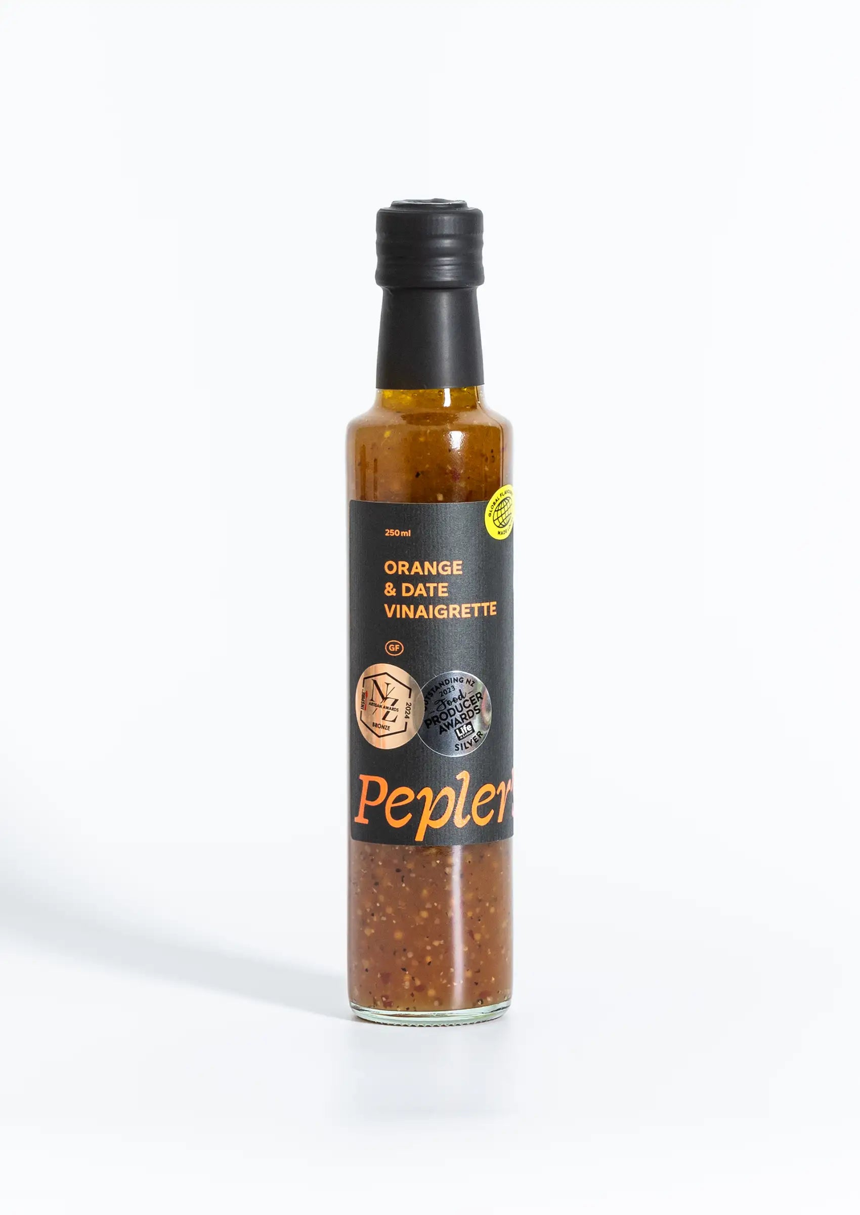 A bottle of Pepler's Orange & Date Vinaigrette sits against a plain white background. The bottle is glass with a black cap and features a label detailing its contents. The main label highlights "Orange & Date Vinaigrette" in bold orange text, with additional information indicating a volume of 250 ml and a gluten-free certification. A silver award sticker is visible on the bottle, showcasing recognition for outstanding products. The vinaigrette has a rich, amber hue, with visible specks of herbs and spices.