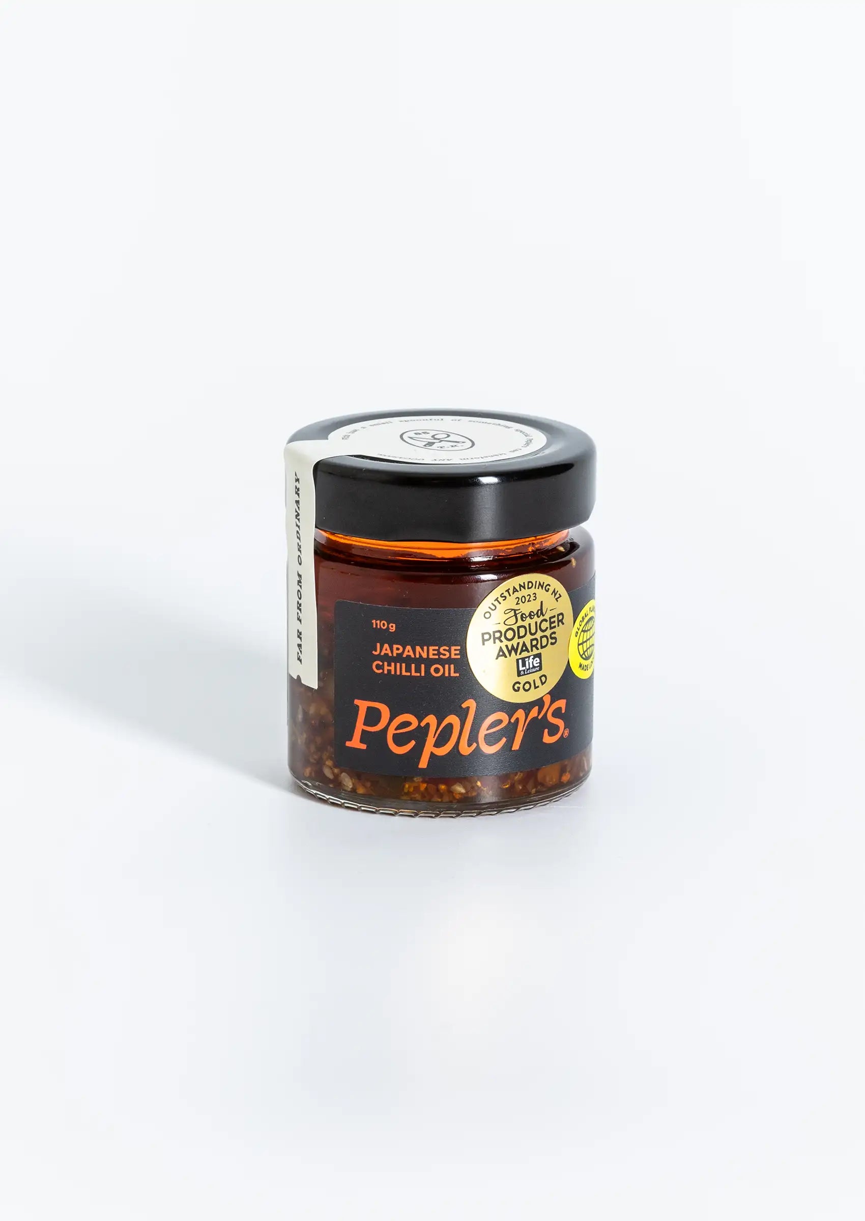 A close-up of a jar of Pepler's Japanese chilli oil, featuring a black lid and a vibrant red and black label. The label displays "110 g" and "JAPANESE CHILLI OIL" prominently, alongside a circular logo that reads "GLOBAL FLAVOURS MADE LOCAL" and another indicating "OUTSTANDING 2021 FOOD PRODUCERS AWARDS GOLD." The jar is set against a soft, neutral background.