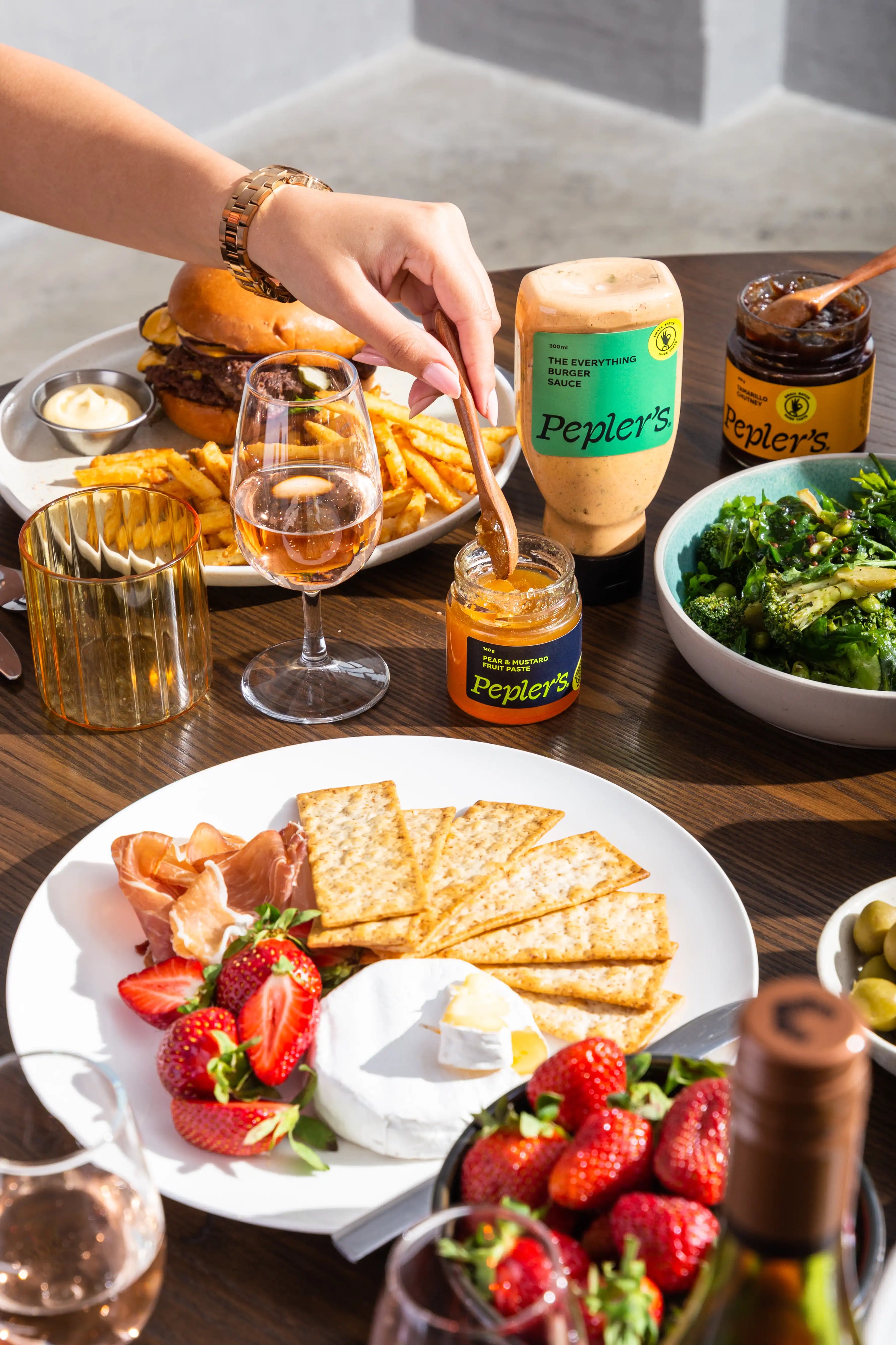 A beautifully arranged table with various foods like a burger, cheese and crackers, broccolini, with a manicured hand dipping a wooden spoon into Pepler's Tamarind & Ginger Chutney. There is also Pepler's Everything Burger Sauce and the Pepler's Tamarind Chutney.