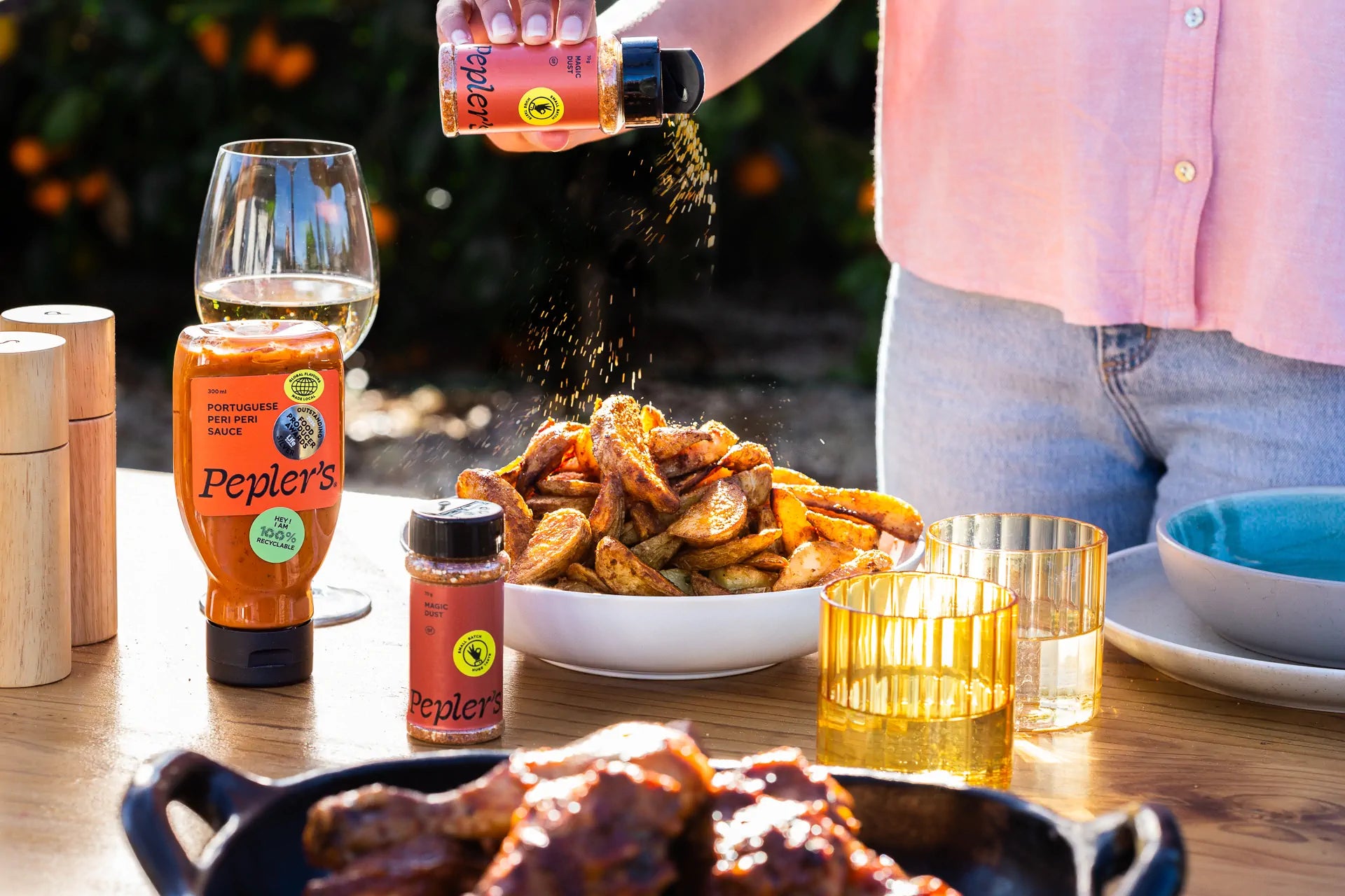 A person in a light pink shirt is sprinkling seasoning over a large bowl of golden, crispy potato wedges on a wooden table. Next to the bowl, there are two bottles of Pepler's sauces, including a Portuguese peri peri sauce and a seasoning labeled "Magic Dust." A glass of white wine and two colourful drinking glasses are also on the table, with a dish of glazed ribs in the foreground and blurred greenery in the background.