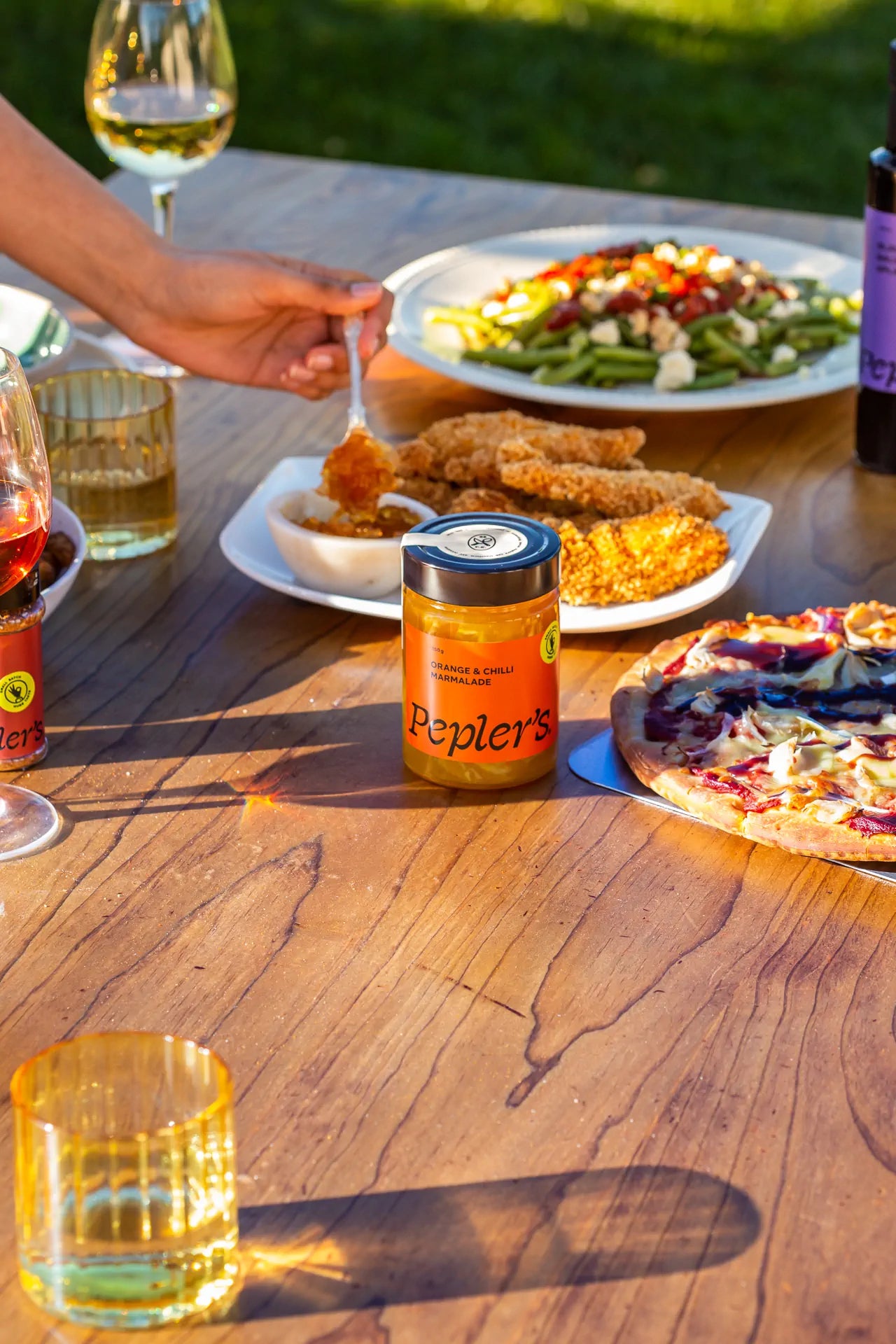 A wooden table is set with various dishes and drinks. In the foreground, a glass of wine and a yellow drink are placed near a jar of orange and chili marmalade labeled "Pepler's." A hand holding a spoon is reaching towards a bowl of the marmalade. To the right, there is a plate with several pieces of fried chicken, while a colourful salad with tomatoes and greens is situated at the back. A pizza with a mix of toppings is also visible on the table, creating a vibrant and inviting dining scene.