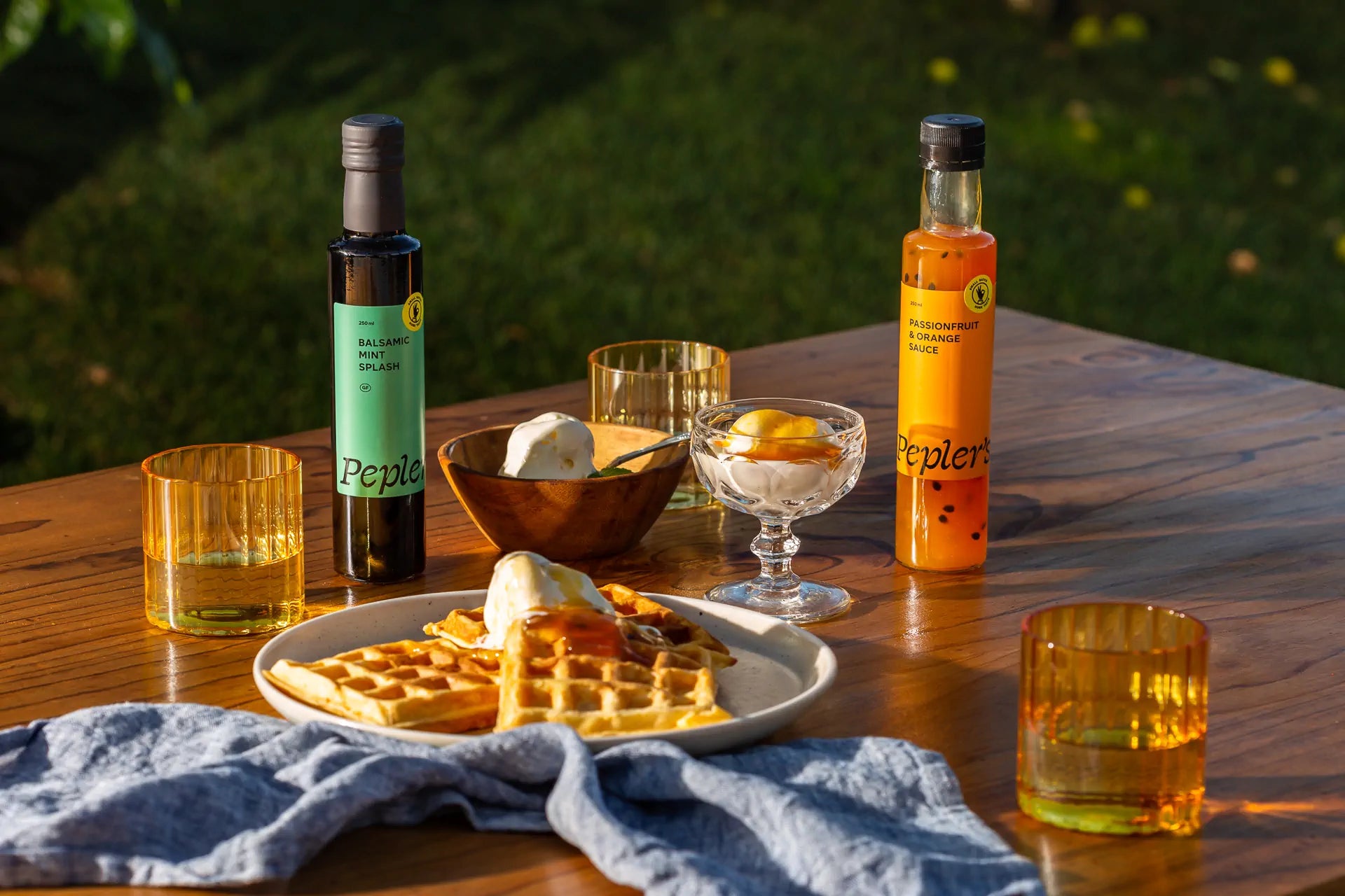 A vibrant outdoor table setting featuring two bottles of Pepler's sauces: a balsamic mint splash and a passionfruit and orange sauce. In the foreground, a plate of golden waffles drizzled with syrup is accompanied by a bowl of ice cream, while a glass dessert dish holds a small portion of fruit. Two amber glasses are on the table, along with a soft blue linen napkin, set against a lush green backdrop with soft sunlight illuminating the scene.